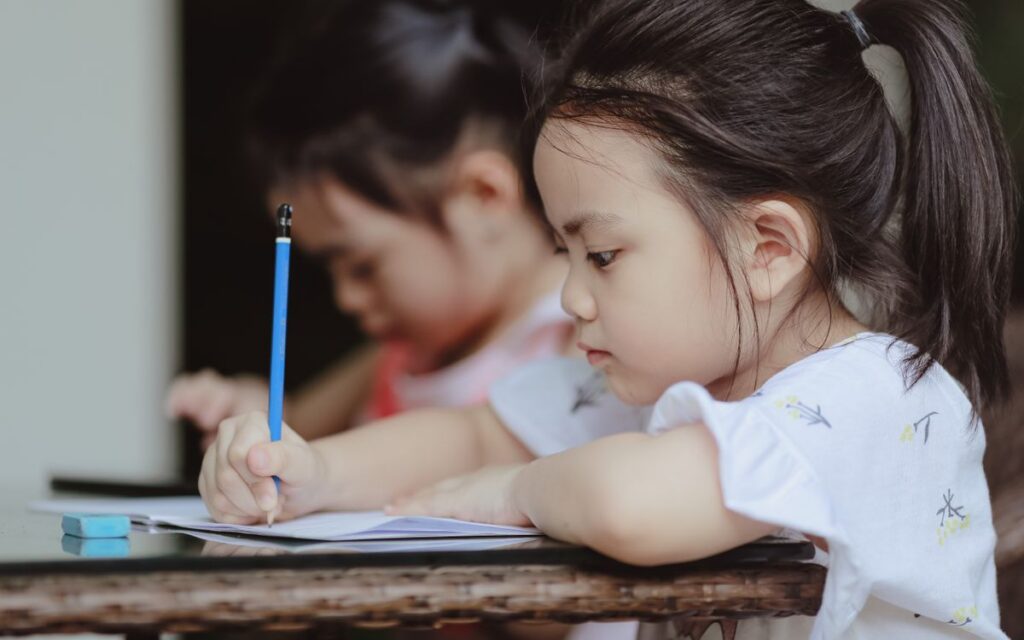 Adorable focused little Asian preschool girls sitting at table and doing tasks in copybooks in classroom