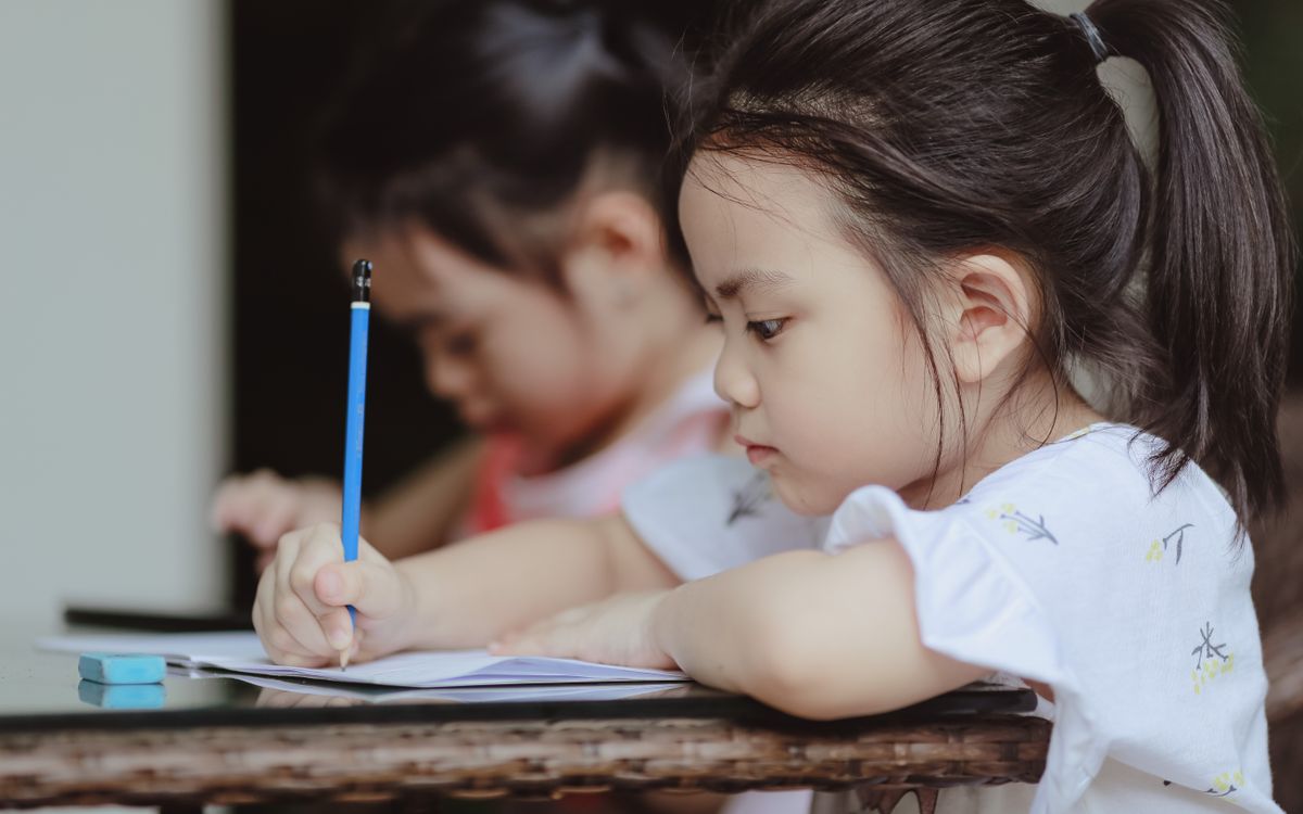 Adorable focused little Asian preschool girls sitting at table and doing tasks in copybooks in classroom