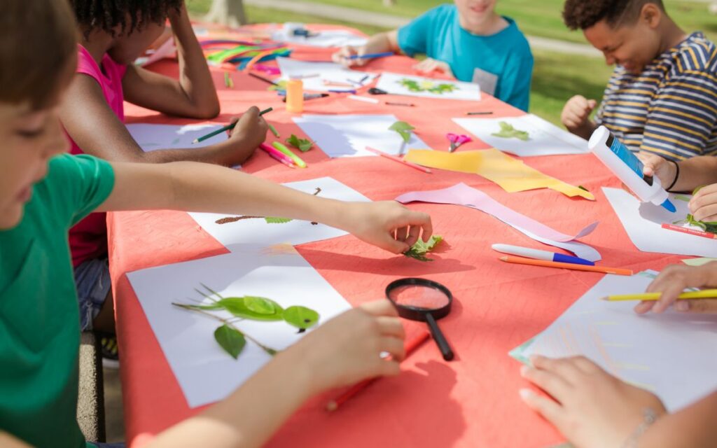 Kids engaging in fun arts and crafts, using leaves and paper outside at a colorful table.