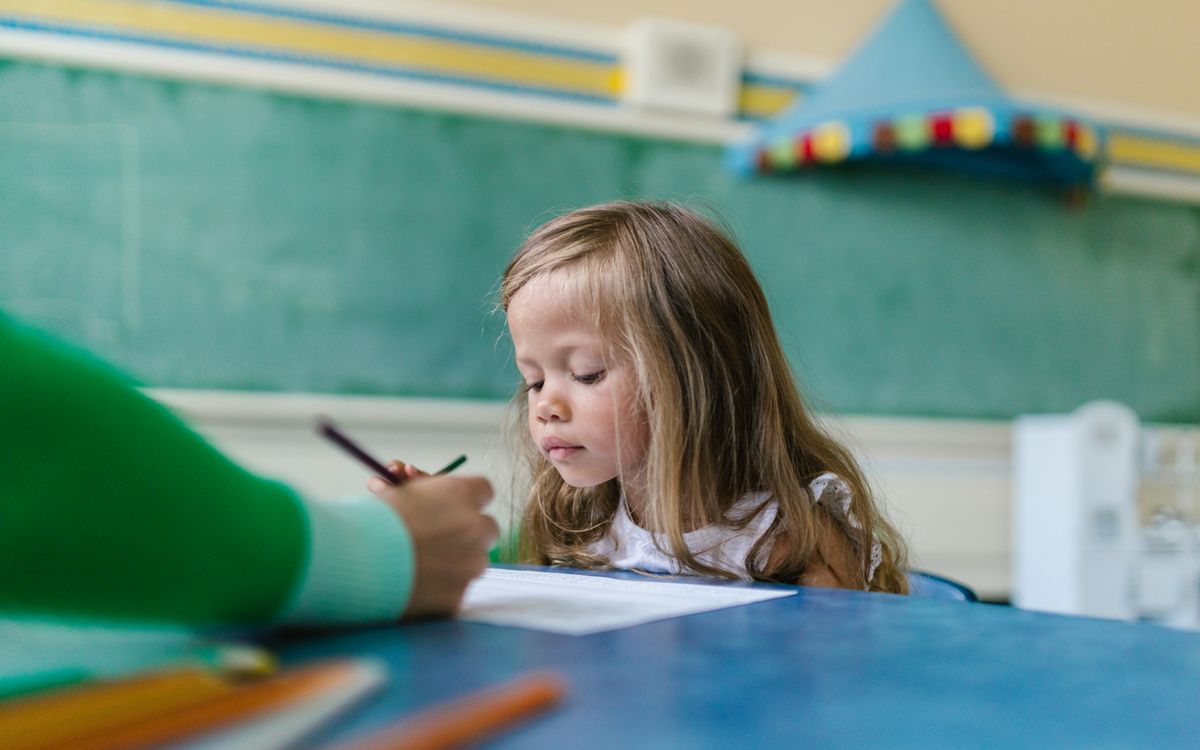 A young girl with long hair concentrates on her drawing in a colorful classroom environment.