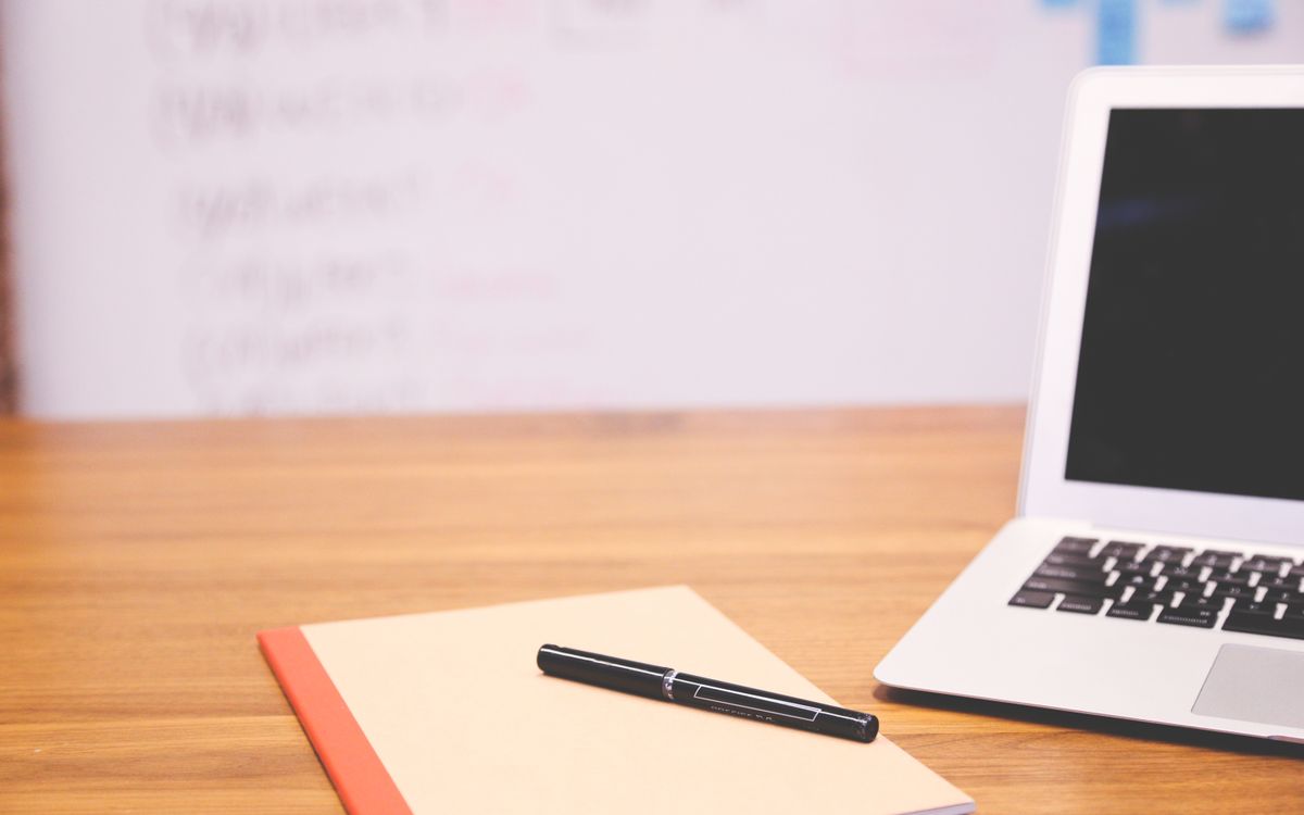 Modern workspace featuring a laptop, notebook, and pen on a wooden desk with a blurred whiteboard in the background.